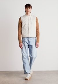 Vesta quilted de un bej deschis, purtată peste un pulover texturat maro, asociată cu blugi de un albastru deschis și adidași în nuanțe neutre, creând o ținută stratificată.