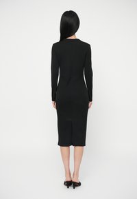 Γυναίκα με μακριά μαύρα μαλλιά, ντυμένη με ένα στενό μαύρο μίντι φόρεμα με μακρυά μανίκια και μαύρες ψηλές πλατφόρμες, που στέκεται γυρισμένη πλάτη σε ένα ουδέτερο φόντο.