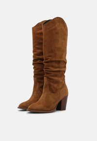 Brązowe zamszowe kozaki za kolano o luźnym kroju, ze szpiczastym noskiem i umiarkowanym, podwyższonym obcasem. Gładka faktura z minimalnymi zdobieniami.