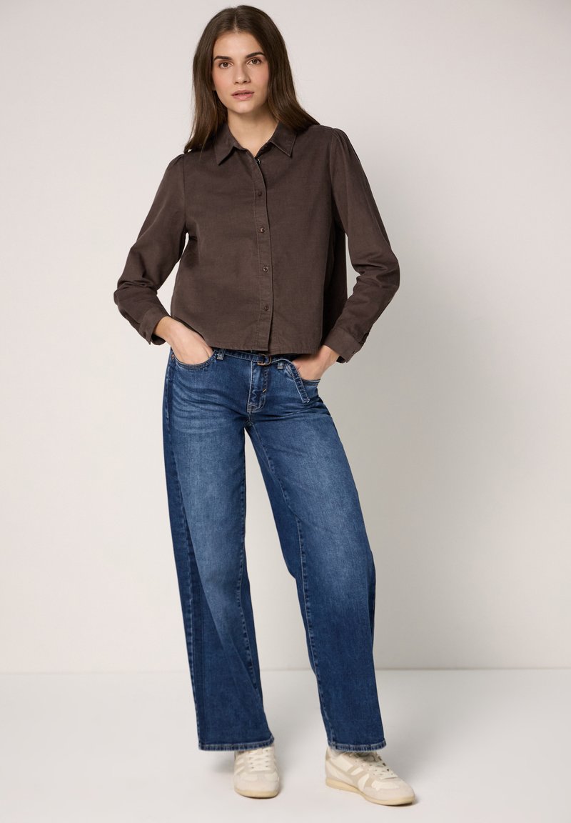 Brown button-up shirt with a collar and long sleeves, paired with wide-leg blue jeans and white sneakers. Smooth fabric and casual fit.