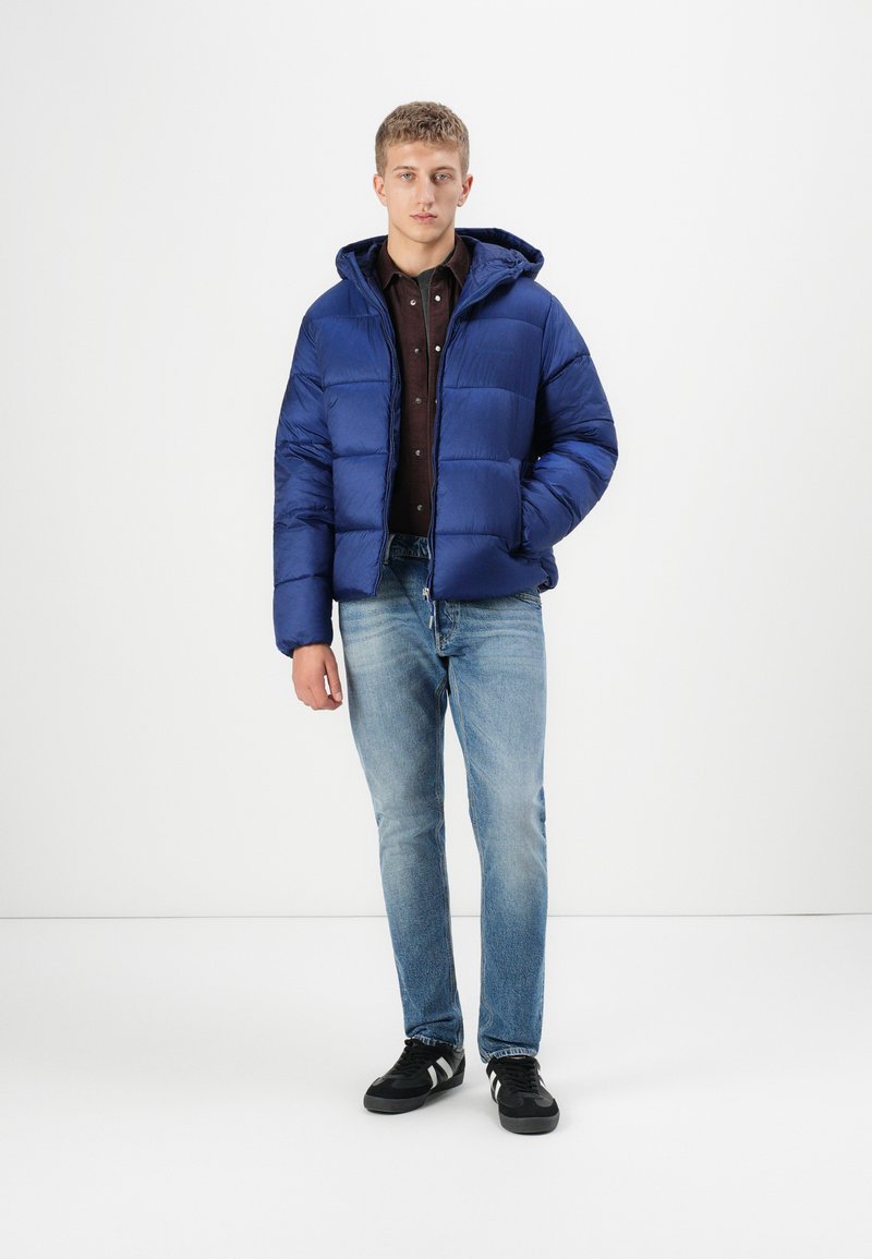 Blaue Pufferjacke mit Kapuze, versehen mit einem gesteppten Design und einem Reißverschluss vorne. Getragen über einem dunklen Shirt, kombiniert mit hellblauen Jeans und schwarzen Sneakers.