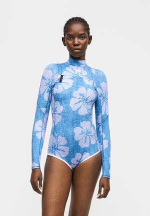 Žena oblečená v modrej dlhoruká neoprenovej kombinéze s veľkými svetlofialovými kvetmi ibišteka, stojaca pred jednofarebným bielym pozadím.