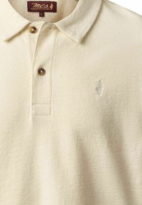 Polo beige avec deux boutons marron, tissu texturé, col classique et logo brodé discret sur le côté gauche de la poitrine.