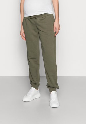 Personne portant un pantalon de survêtement vert olive avec cordon, des baskets blanches et une chemise blanche, debout devant un fond uni.