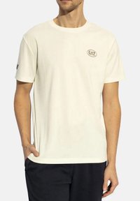 T-shirt in cotone crema chiaro con maniche corte, scollatura rotonda e un piccolo logo ovale sul petto. Vestibilità dritta e texture morbida.