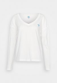 Chemise blanche à manches longues en V avec un logo en forme de cœur bleu sur la poitrine gauche, présentée sur un fond gris clair uni.