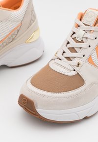 Baskets beige clair et marron clair avec lacets blancs, empiècements en mesh et en daim, accents orange et semelles blanches épaisses sur fond blanc.