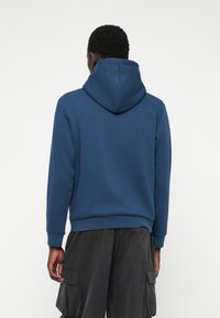 Hoodijs tums zila auduma ar mīkstu tekstūru. Aprīkots ar kangaru kabatu un ribotām piedurknēm, valkāts ar tumšiem kravas biksēm.