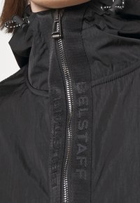 Chaqueta ligera negra con un cuello alto, cuenta con un cierre de cremallera y un ribete de tejido con marca. Textura suave y detalles de diseño mínimos.