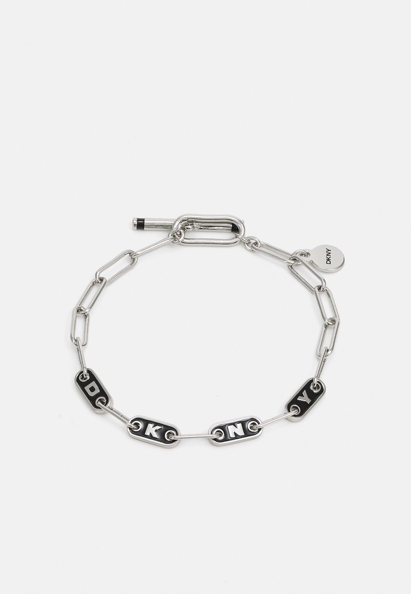Bracelet chaîne en argent avec des maillons rectangulaires noirs formant "DKNY" et un pendentif rond gravé "DKNY" sur fond blanc.