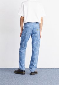 Blaue Denim-Cargo-Hosen mit lockerer Passform, die mehrere Taschen und ein wiederholtes Logomuster aufweisen. Kombiniert mit einem schlichten weißen Hemd und schwarzen Schuhen.