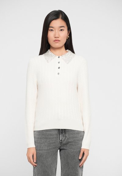Jeune femme portant un pull en maille torsadée blanche avec un col perlé et des boutons argentés, associé à un jean en denim gris.