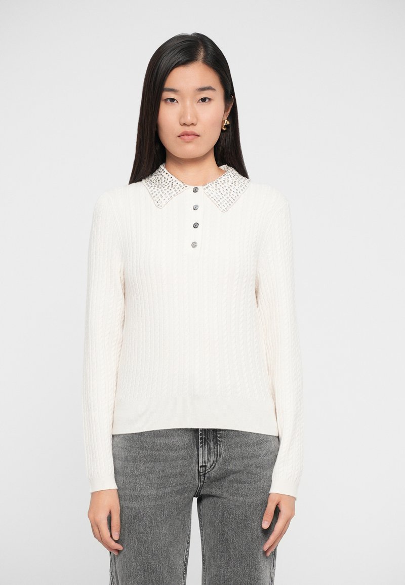 Ung kvinde iført en hvid kabelstriktrøje med en perlekant og sølvknapper, sammen med grå denimjeans.