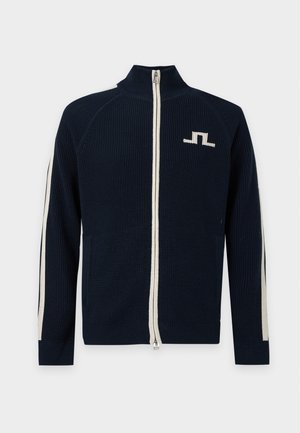 Jeune homme portant une veste bleu marine zippée avec des bandes blanches sur les manches, un pantalon beige et des chaussures habillées noires, debout devant un fond uni.