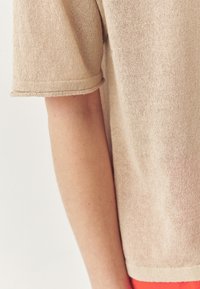 Top beige lavorato a maglia con maniche corte arrotolate, che presenta un tessuto testurizzato e una vestibilità ampia, abbinato a pantaloni rossi vivaci.