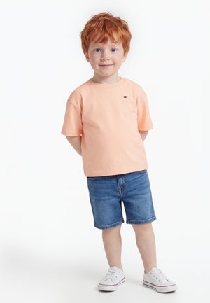 Jeune garçon roux debout, mains derrière le dos, portant un t-shirt pêche, un short en jean et des baskets blanches sur fond blanc.