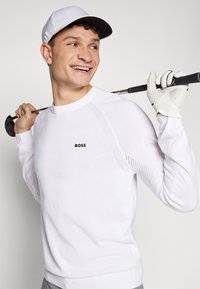 Valge golfisweater, millel on õlgadel hingavad võrgukujundused, töötleva logo ja soonikdad varrukad. Kanda koos kindaga ja mütsiga.