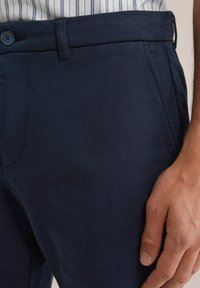 Detailní záběr na osobu v námořnických kalhotách s knoflíkem a poutkem na pásek a pruhované košili zastrčené dovnitř.