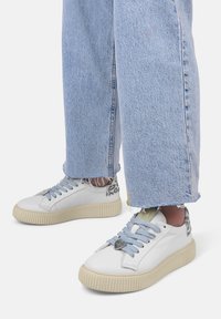 Sneakers bianchi in pelle con suola beige texturizzata, lacci blu e dettagli in stampa leopardata. Indossati con jeans chiari strappati.
