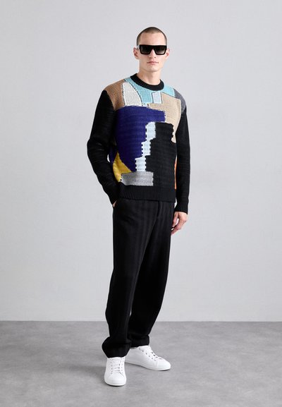 Pull tissé multicolore avec des motifs géométriques, manches en tricot noires, associé à un pantalon noir texturé et des baskets blanches.