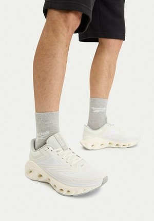 Gambe con pantaloncini neri, calzini Reebok grigio chiaro e sneaker Reebok bianche con suole spesse e perforate su sfondo bianco.