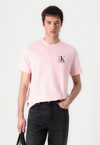 T-shirt en coton rose clair à manches courtes, col rond, avec un petit logo noir Calvin Klein sur la poitrine gauche. Porté avec un jean noir.