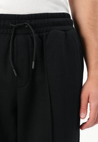 Primer plano de la cintura de una persona que lleva pantalones de chándal negros con cordón y una camiseta blanca, con la mano derecha relajada a su lado.