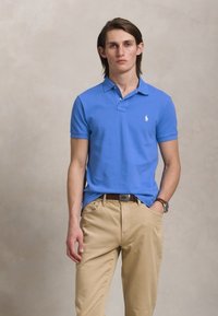 Spilgtas zilas kokvilnas polo krekls ar īsām piedurknēm un apkakli. Beigžu krāsas chinos bikses un brūna josta. Modelis stāv uz neitrālas fona.