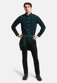 Groene en marineblauwe geruite overhemd met lange mouwen, gecombineerd met zwarte broek. Een groene hoodie is om de taille geknoopt. Zwarte schoeisel.