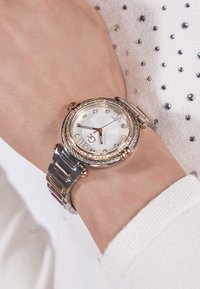 Hõbe- ja kuldtooni randmekell, millel on ümar nägu, valge kelladial ja teemantmarkerid, ning linkidega metallist rihm tekstureeritud viimistlusega.