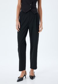 Schwarze maßgeschneiderte Hose mit geradem Schnitt und Falten, aus einem glatten Stoff gefertigt. Kombination mit schwarzen Lackleder-Slingback-Absätzen.