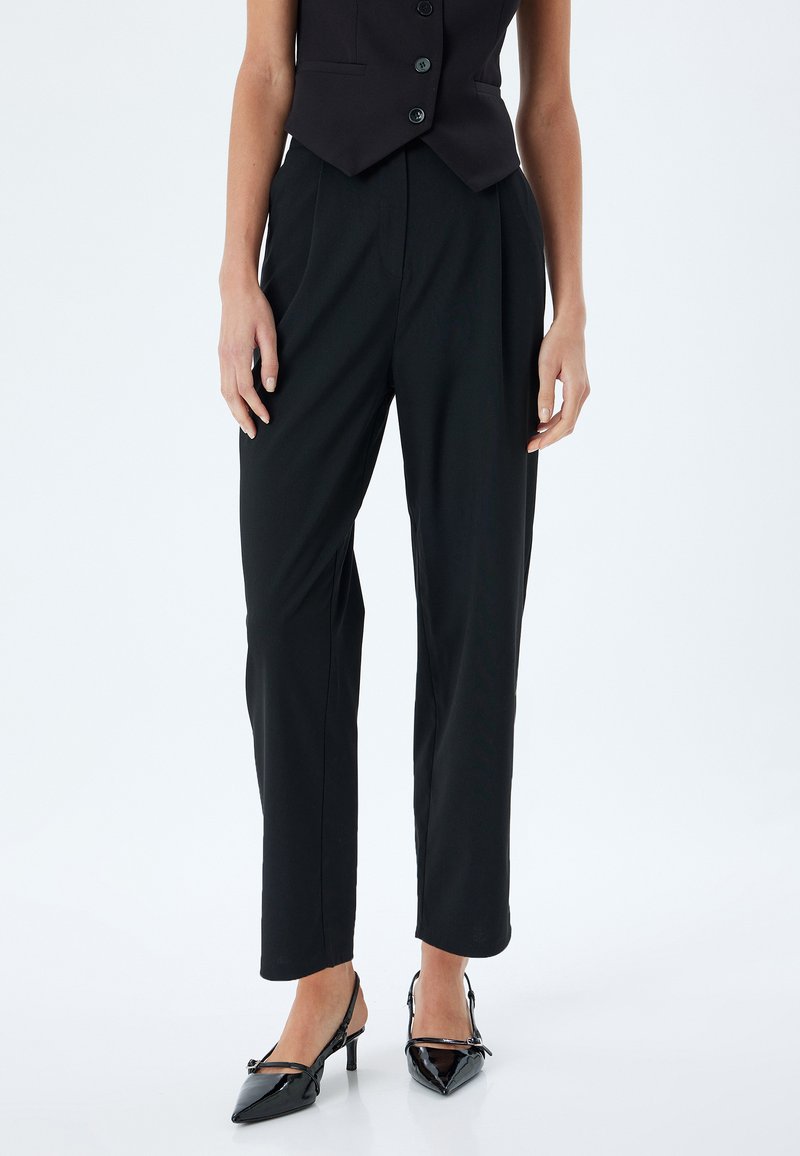 Schwarze maßgeschneiderte Hose mit geradem Schnitt und Falten, aus einem glatten Stoff gefertigt. Kombination mit schwarzen Lackleder-Slingback-Absätzen.