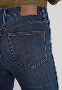 Dark blue denim jeans feature a back pocket with subtle stitching details and a brown leather patch displaying the brand name.