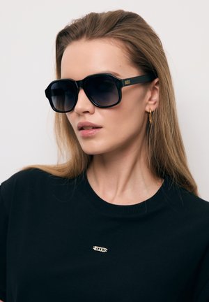 Kobieta z długimi blond włosami, nosząca duże czarne okulary przeciwsłoneczne, złote kolczyki koła oraz czarny top z małym złotym logo na piersi.