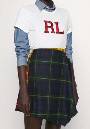 Personne porte un T-shirt blanc avec les lettres rouges "RL" sur une chemise en jean bleu à boutons, associé à une jupe asymétrique à carreaux verts et jaunes.