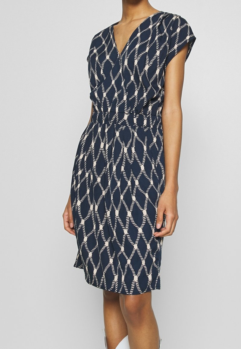 Robe bleu marine avec un motif géométrique crème, manches courtes, encolure en V et taille élastique. Tissu léger, longueur genoux.