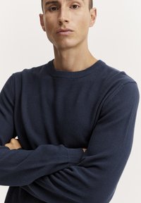 Tamsiai mėlynas struktūrizuotas megztinis su apvalia iškirpte ir raštuotais rankogaliais, pasižymintis tolygiu raštu ir laisvu siluetu.