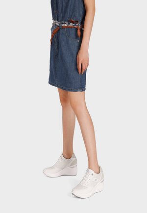 Donna che indossa un vestito di jeans al ginocchio con cintura decorata e sneakers bianche con platform, in piedi davanti a uno sfondo semplice.