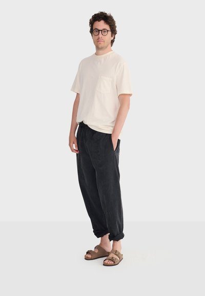 Homme aux cheveux bouclés et portant des lunettes, vêtu d'un t-shirt beige à poche, d'un pantalon noir ample retroussé aux chevilles et de sandales marron à double bride, debout.
