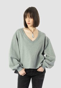 Felpa verde chiaro, oversize, con scollo a V e maniche lunghe e voluminose. Abbinata a pantaloni neri e una collana con pendente argento.