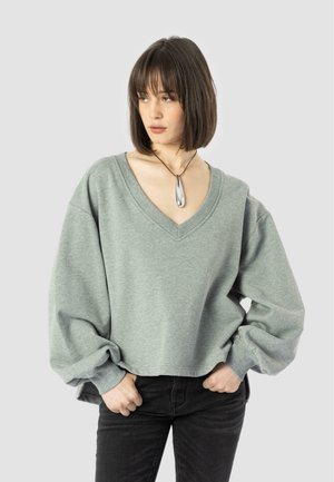 Light green, oversized, V-neck sweatshirt with long, voluminous sleeves. Paired with black pants and a silver pendant necklace.