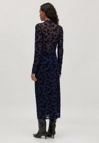 Ein langes navyblaues Kleid aus durchscheinendem Stoff mit floralem Muster, das lange Ärmel und eine taillierte Silhouette aufweist. Kombiniert mit schwarzen spitzen Stiefeln.