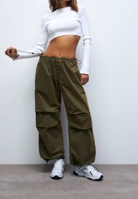 Pantalon cargo vert olive avec une taille à cordon de serrage, doté de plusieurs grandes poches et d'une coupe ample. Associé à un haut blanc à manches longues court.