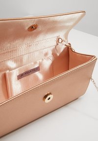 Kabelka v růžovém zlatě se hladkým koženým vnějškem, s saténově podšívkou uvnitř a zlatým kruhovým zapínáním na klapku. Obsahuje odnímatelný řetízkový popruh.
