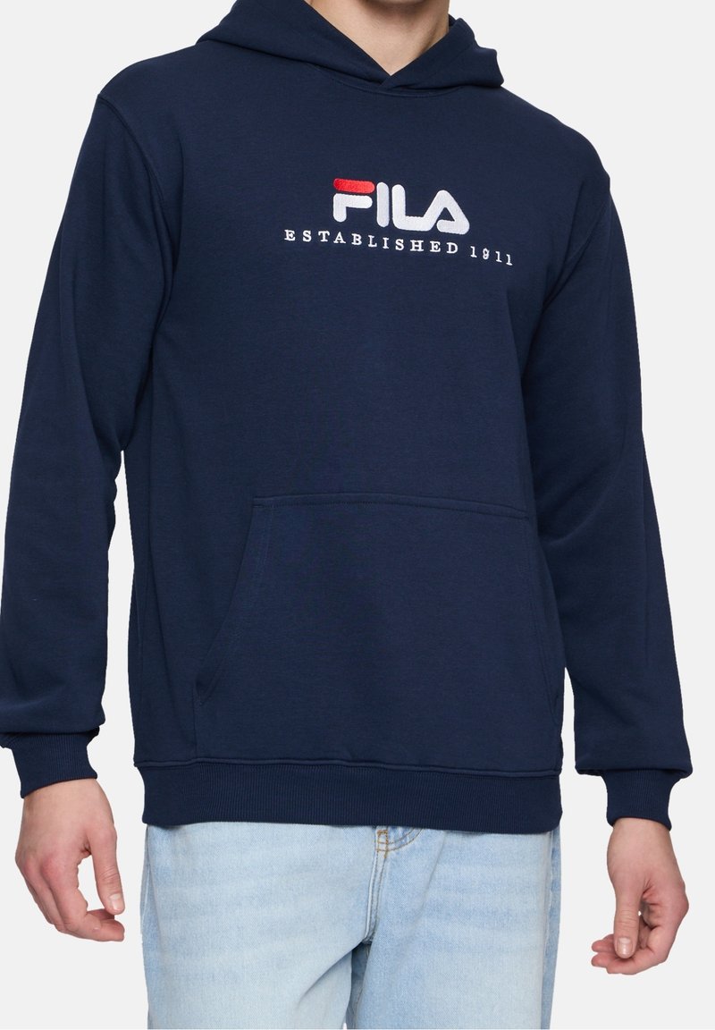 Sweat à capuche bleu marine avec poche avant, logo « FILA Established 1911 » rouge et blanc sur la poitrine, porté par une personne en jean bleu clair.