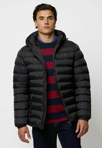 Schwarze Pufferjacke mit Kapuze und gestepptem Design, ausgestattet mit einem Frontreißverschluss und seitlichen Taschen. Trägt eine gestreifte Rundhals-Sweatshirt darunter.