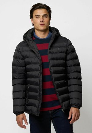 Jachetă puffer neagră cu glugă și design matlasat, având un fermoar frontal și buzunare laterale. Stratificată peste un pulover cu guler rotund în dungi.