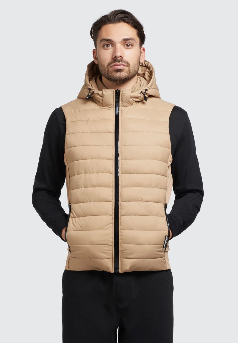 Tan puffervest met een capuchon, voorzien van horizontale quilttextuur, een ritssluiting aan de voorkant en zijzakken. Gemaakt over een zwart shirt met lange mouwen.