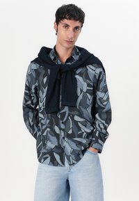 Knappskjorte med et mørkeblått bladmønster, sammen med en strikket marineblå genser knyttet rundt skuldrene. Lys blå jeans.