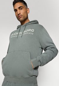 Szürke kapucnis pulóver, elülső zsebbel, bordázott mandzsettával, a mellkason fehér felirattal: "BJÖRN BORG TRAINING CLUB MONACO".